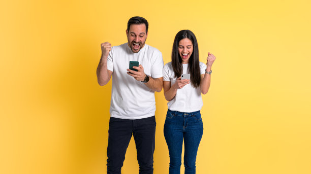 兴奋的年轻夫妇穿着白色t恤，一边欣喜若狂地尖叫，一边挥舞着拳头，一边用黄色背景隔离的手机阅读好消息图片下载