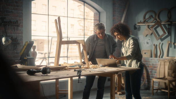 家具车间的小企业主使用笔记本电脑和讨论新木椅的设计。中年木匠和年轻的女学徒在阁楼工作室工作。图片下载