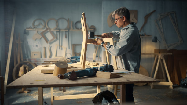 中年木匠戴上眼镜，阅读蓝图，开始组装木椅的零件。时尚家具设计师在阁楼空间的工作室工作，墙上有工具。图片下载