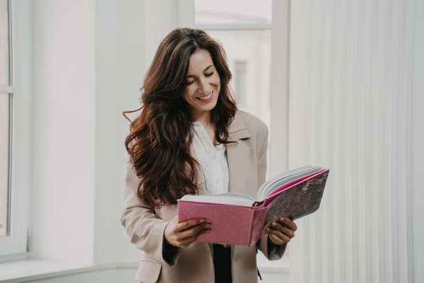开朗时尚的褐发女人在米色夹克抱着书读着露齿的微笑在家里。迷人的西班牙女商人在办公室笑得很开心。成功的人。满足女性。图片下载
