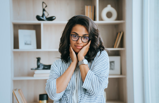 可爱的非裔美国年轻女商人在衬衫和眼镜站在家里靠书架看镜头触摸脸满意的护肤霜。满意的巴西女孩后水疗程序。图片下载