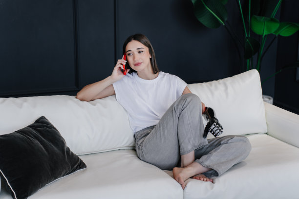 可爱的黑发学生女孩残疾打电话看镜头，露齿微笑，在白色t恤和牛仔裤坐在舒适的沙发上。意大利女孩戴着人工仿生假肢。图片下载