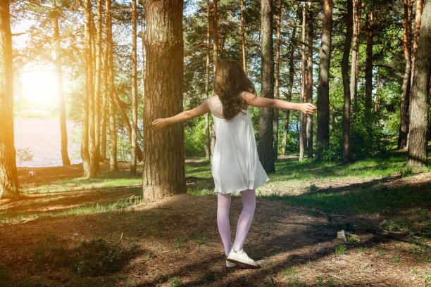 后视图小女孩在白色连衣裙双臂两侧在绿色森林户外，转身离去。完整的长度女孩在阳光林地摆姿势的背影。环保理念。本空间图片下载