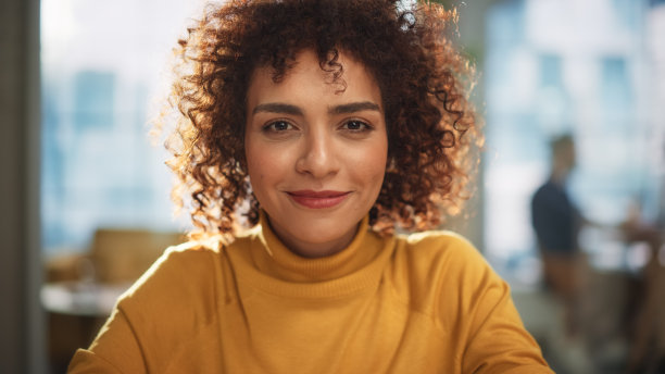 一位迷人的阿拉伯女性坐在创意机构的办公桌前。年轻时尚的经理与卷发微笑，看着相机。在现代公司工作的经理。图片下载