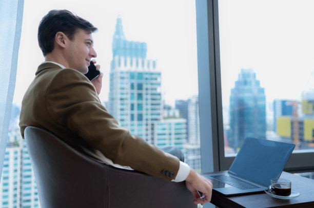 年轻的商人坐在窗边的放松室里，俯瞰美丽的城市建筑。一起打电话谈生意。图片下载