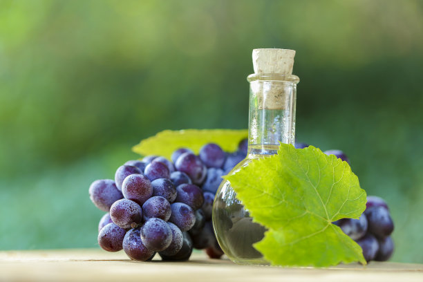葡萄醋在玻璃圆瓶，葡萄串与绿叶在一个木锯切割在模糊的绿色花园背景。有机天然生物葡萄籽油。图片下载