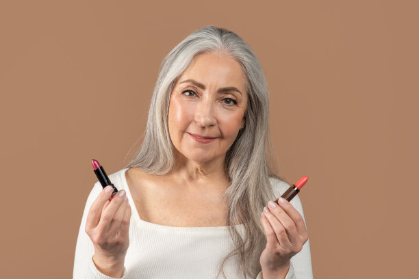 美丽的成熟女子手持两支口红的肖像，在棕色的工作室背景上选择最好的颜色图片下载
