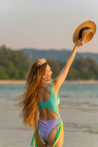 一个美丽的白人女人在比基尼性感的背影在海滩上拿着一顶沙滩帽。女士享受海风和日落美景图片下载