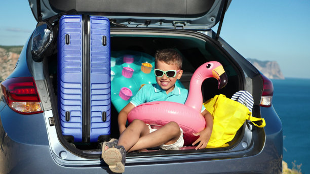 兴高采烈的男孩戴着墨镜坐在后备箱里，拿着一个旅行箱，一个充气圈。图片下载