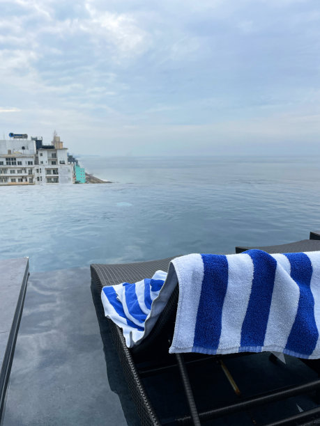 蓝色和白色条纹毛巾留在编织塑料躺椅上的图像，无边泳池俯瞰科伦坡的城市景观，斯里兰卡，蓝色多云的天空背景，重点在前景图片下载