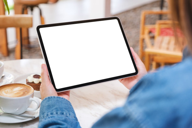 在咖啡馆的桌子上，一个女人拿着空白白色桌面屏幕的数字平板电脑和蛋糕和咖啡杯的模型图像图片下载