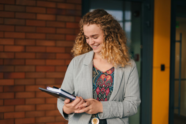 一个快乐的卷发女学生手持书籍走出校园的肖像，并在户外相机前摆姿势。准备学院和大学的概念。现代的生活方式。现代成功人士。快乐的生活方式。图片下载