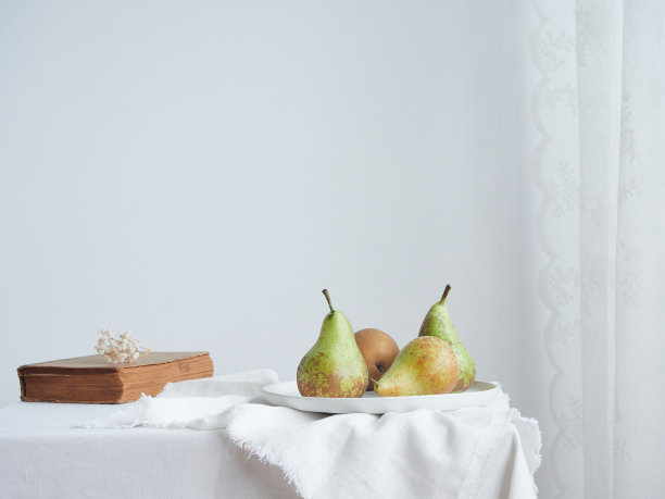简单极简的室内生活方式构成:梨子放在陶瓷盘上，旁边是一本旧书，上面放着干花，桌子上有白色桌布，旁边是网帘。背景中带有复制空间的浅色墙壁。图片下载