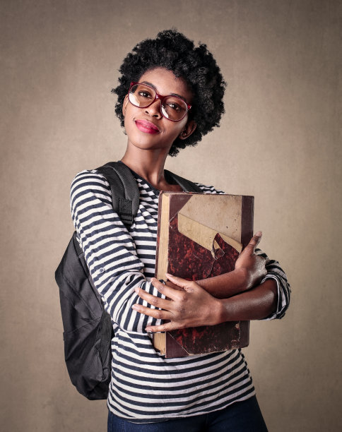 一个年轻的女学生，戴着眼镜，背着书包，手里拿着一本大书图片下载