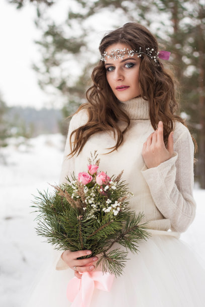 在白雪覆盖的冬季森林里，美丽的新娘身着白色婚纱，手捧花束。新娘的自然肖像图片下载