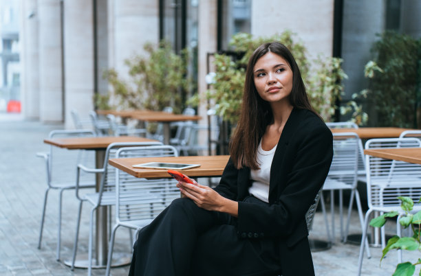 不开心的韩国黑发女商人坐在咖啡馆外面拿着电话，看着一边等待伙伴。疲惫的年轻女律师打字信息。漂亮的亚洲女孩在等男朋友。经理。图片下载