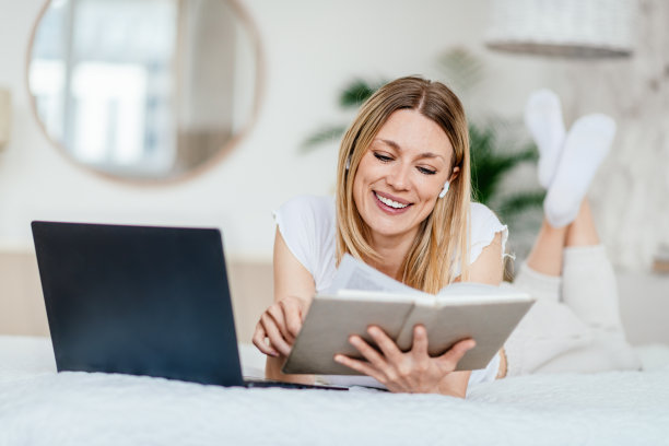 微笑年轻的欧洲金发女性在无线耳机看书或笔记本，躺在床上图片下载