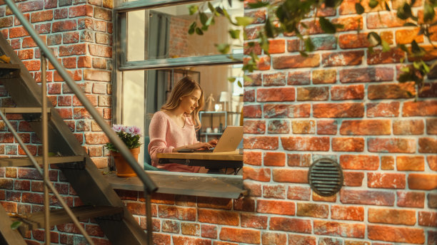 一个年轻女子坐在多层砖房的家里，用笔记本电脑工作的肖像。积极主动的女性在家工作时保持微笑和快乐。图片下载