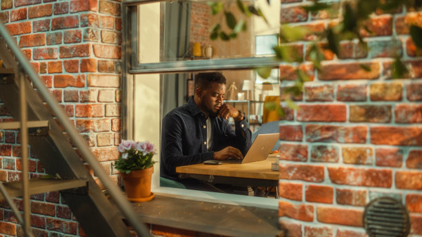 年轻的黑人在家里使用笔记本电脑。白天做一些鼓舞人心的远程工作。专业从事计算机工作，撰写创意项目计划。从窗外拍摄。图片下载