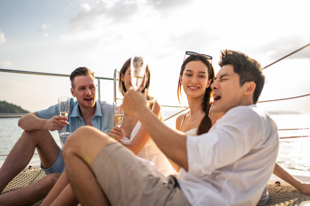 一群不同的朋友在游艇上喝香槟聚会。迷人的年轻男人和女人挂在一起，庆祝假期度假旅行，而双体船航行在夏天日落。图片下载