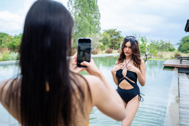 亚洲女性朋友穿着比基尼在游泳池用手机拍照。迷人的年轻性感的女孩闲逛，庆祝暑假享受阳光明媚的日子户外旅行假期在一个私人别墅。图片下载