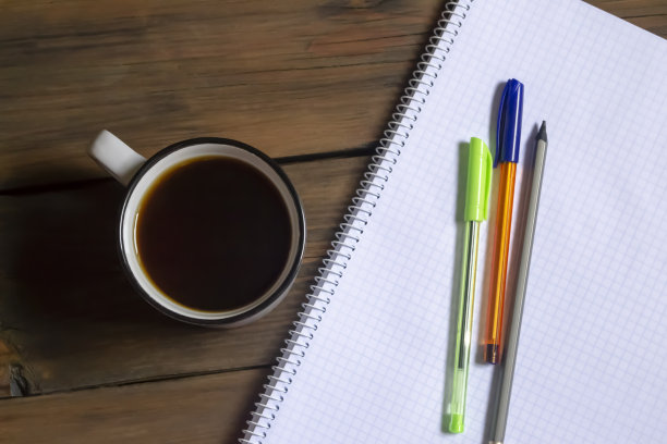 笔记本和笔旁边放着一杯咖啡。桌子上有一本格子笔记本，三支钢笔和一杯黑咖啡。在他学习的时候。图片下载