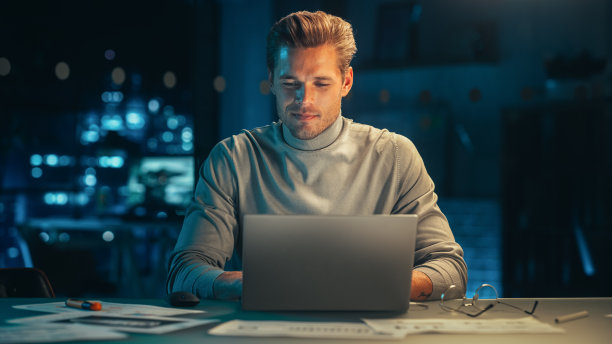 晚上在公司办公室里摆弄笔记本电脑的帅哥。分析报告的年轻专家。营销助理为客户制定消费者销售策略。图片下载