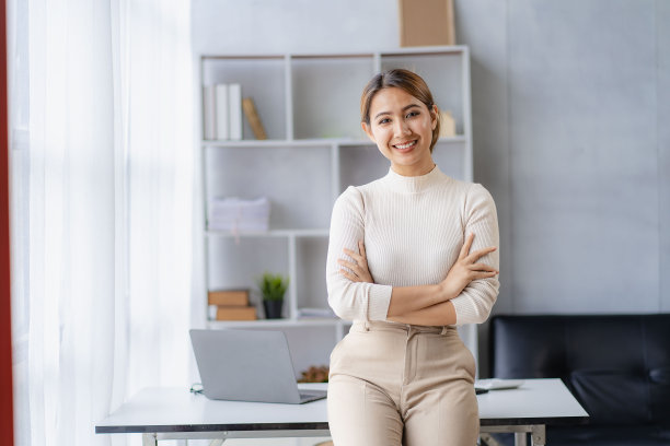 年轻的亚洲女性双手交叉站在他的桌子前，背景是笔记本电脑。图片下载