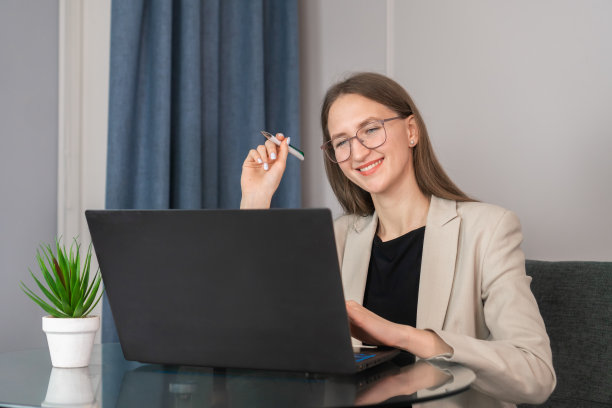 年轻的女商人，穿着夹克，戴着眼镜，面带微笑，头边拿着一支笔，坐在室内的玻璃桌旁，看着笔记本电脑。事业理念成就，自由职业图片下载