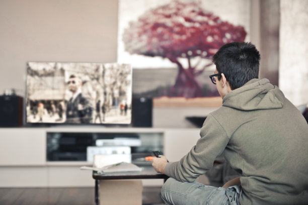 一个戴着眼镜，穿着灰色连帽衫的男孩正在看电视，背景是一棵红色的树图片下载