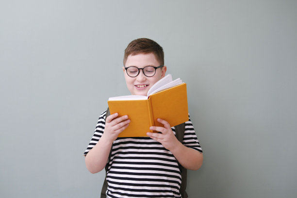 一个可爱开朗的男孩，穿着带玻璃的条纹t恤，正在阅读一本黄色的书，背上有绿色背景的书包。图片下载