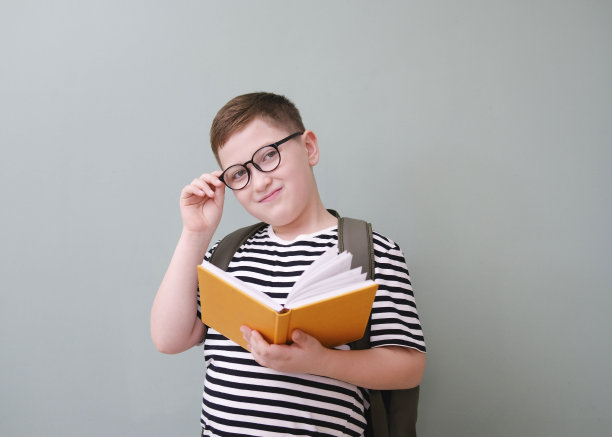 一个可爱开朗的男孩，穿着条纹t恤，摸着眼镜，在绿色背景上拿着一本黄色的书和书包。图片下载