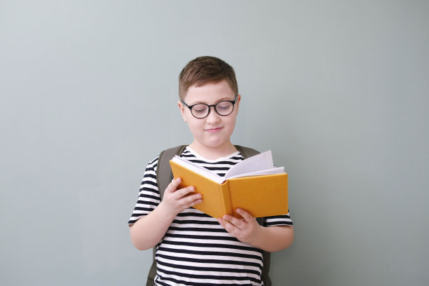 一个可爱开朗的男孩，穿着带玻璃的条纹t恤，在绿色背景上拿着一本黄色的书和书包。图片下载