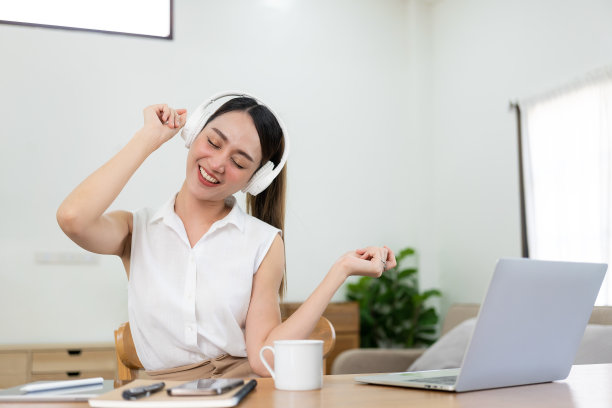 漂亮的亚洲女孩戴着耳机在笔记本电脑和手机上开心地听音乐。享受假期在工作时听音乐，在家办公的概念。图片下载