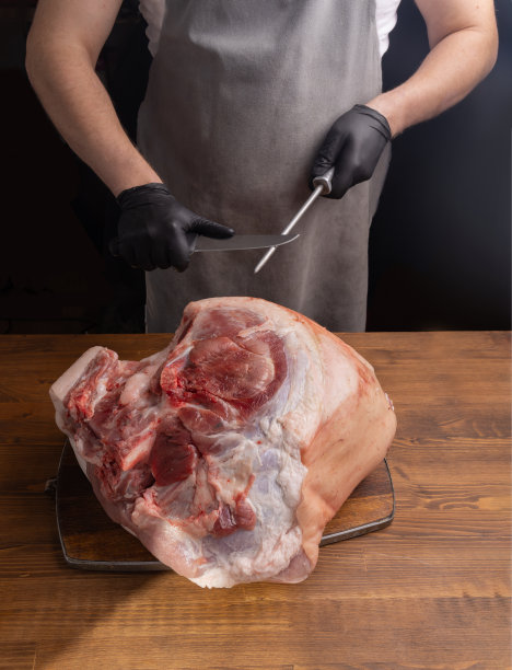 一个穿灰色围裙的人正在磨一把肉刀。桌上放着一块生肉。切肉用的刀。图片下载