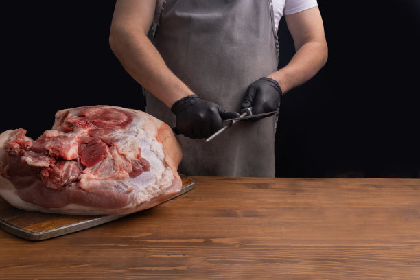 一个穿灰色围裙的人正在磨一把肉刀。桌上放着一块生肉。切肉用的刀。图片下载