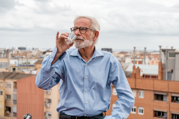 成熟的商人，灰色头发和胡子，穿着蓝色衬衫和牛仔裤，戴着眼镜在屋顶露台上喝咖啡，背景是城市天窗。本空间图片下载