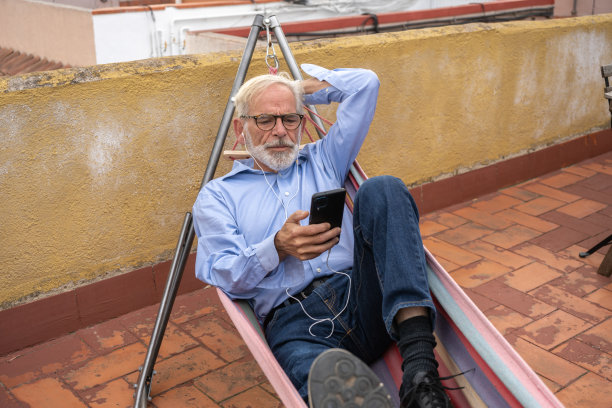成熟的商人，灰色头发和胡子，蓝色衬衫和牛仔裤躺在吊床上，智能手机在阳台上图片下载