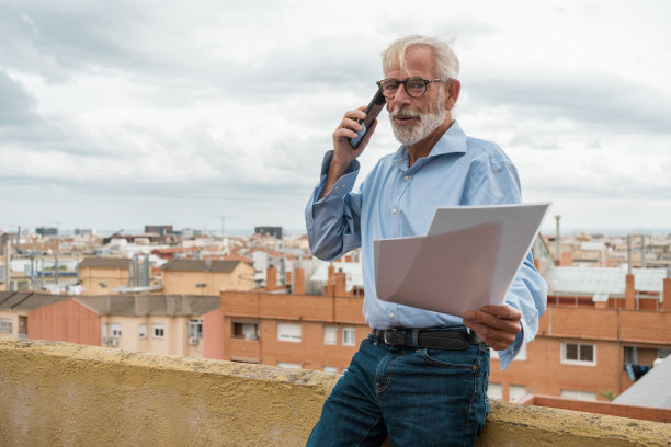 成熟的商人，灰色头发和胡子，蓝色衬衫，牛仔裤和眼镜，纸质文件和智能手机在屋顶露台上，背景是城市天窗。本空间图片下载