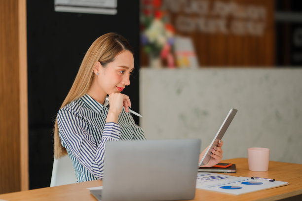 在明亮现代的办公室里，微笑的亚洲女商人在她的办公桌上用平板电脑工作，用图表文件计算财务报告费用图片下载