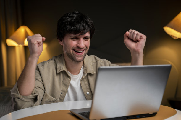 成功和成就的概念。一个男人看着笔记本电脑面带幸福开朗的笑容，实现人生目标。一个快乐的男人坐在卧室里做一个胜利的手势。图片下载