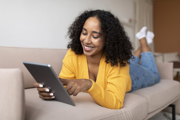 放松的年轻黑人女子躺在沙发上，使用平板电脑，上网聊天或查看社交媒体图片下载
