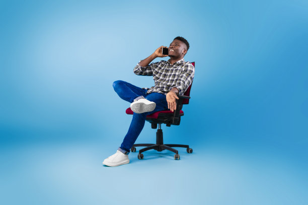 快乐的年轻黑人男子打电话，坐在椅子上，在蓝色的演播室背景上进行电话交谈图片下载