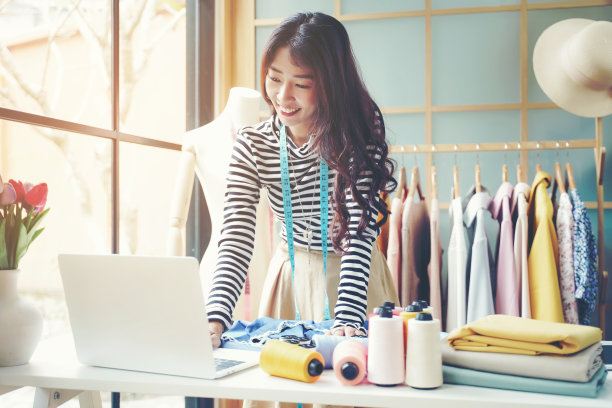 年轻的时装设计师使用笔记本电脑工作，并在展厅接受设计裁缝和布料的订单客户，图片下载