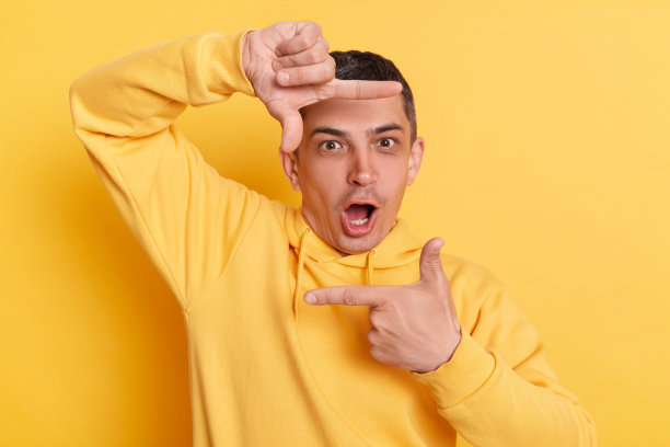 震惊了摄影师。惊讶惊讶的男子穿着休闲帽衫，手指框架看着相机与张开的嘴摆孤立的黄色背景。图片下载