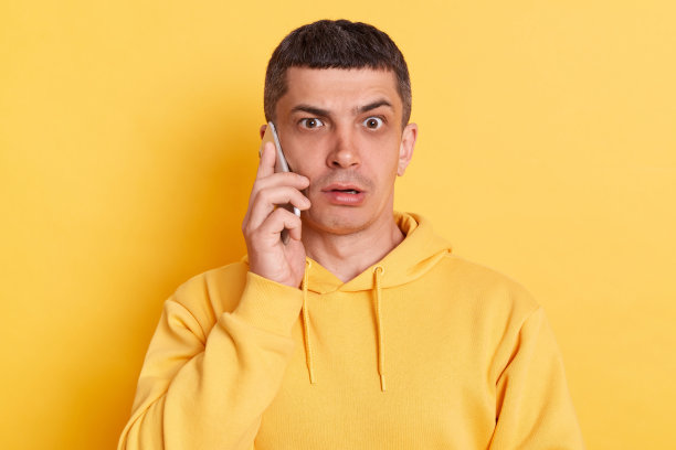 一名穿着休闲帽衫的男子在智能手机上说话，听到令人惊讶的消息，大眼睛看着相机，在黄色背景上摆出孤立的姿势。图片下载