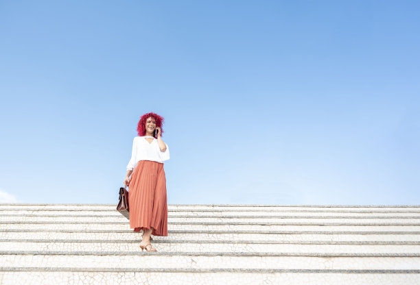 一个快乐的时尚女商人的水平肖像，红色爆炸头的头发拿着手提包，走下白色的楼梯，在蓝天的背景下讲智能手机图片下载