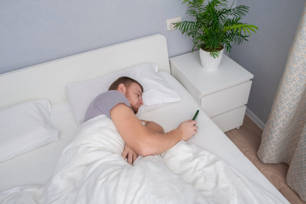 留胡子的男人在家里的床上打电话。不用起床就能购物。人们对电子产品的依赖。网络成瘾。对手机的依赖小玩意上瘾图片下载