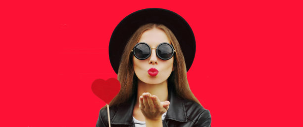 时尚的年轻女子的肖像与红色心形棒棒糖吹她的嘴唇送甜蜜的飞吻，戴着黑色圆形帽子的背景图片下载
