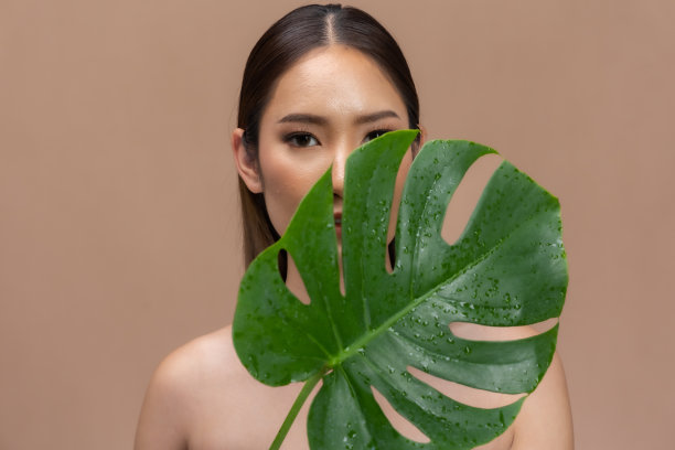 美丽的亚洲女人动人的眉毛微笑，干净和新鲜的皮肤在米色背景上幸福..美丽的肖像，迷人的年轻女孩与绿色的植物叶子隔离。图片下载